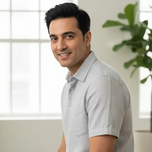 Professional headshot of a smiling man with dark hair, wearing a light gray short-sleeve collared shirt, seated indoors next to a large window and a potted plant.